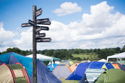 a landscape photo of a post with several different signs directing to the areas of the festival which is towering above fields of pitched tents